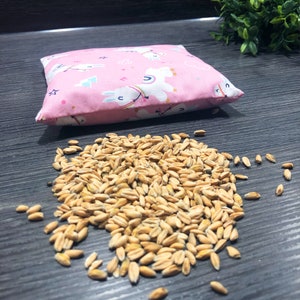 Könnte beinhalten: Ein rosa Stoffbeutel mit einem weißen Einhornmuster und einem Haufen Weizenkörner auf einer dunklen Holzoberfläche.