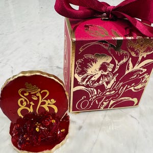 May include: A red and gold decorative box with a floral design and the text "Especially for you". The box is tied with a red ribbon. A small red and gold figurine of the Hindu god Ganesha is sitting on a white surface.