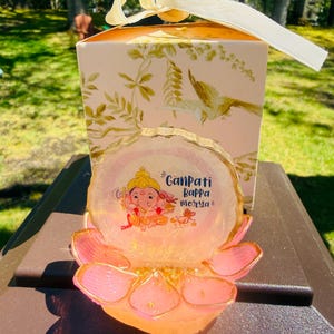 May include: A pink and gold lotus flower-shaped candle holder with a clear, round top featuring an illustration of a deity and the text "Ganpati Bappa Morya". A matching gift box with a cream ribbon is in the background.