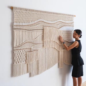 Op de afbeelding: Grote, beige macramé wandkleed met een houten staaf. De textielkunst toont verschillende geweven patronen, waaronder knopen en franjes, waardoor een gestructureerd, gelaagd ontwerp ontstaat. Het kunstwerk wordt aan een witte muur getoond.