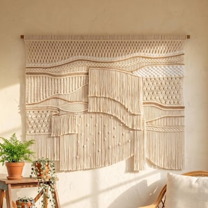 May include: A large, beige macrame wall hanging with various woven patterns hangs above a wicker chair with a cream cushion. A wooden ladder holds potted plants, and a copper watering can sits nearby, creating a cozy, bohemian-style interior.