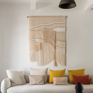 May include: A white couch with multiple pillows in a living room. The couch has a wooden frame and a white fabric cover. The pillows are in various colors and textures, including beige, yellow, and red. A macrame wall hanging is on the wall behind the couch. The wall hanging is made of natural fibers and has a geometric design. A black side table is in the corner of the room.