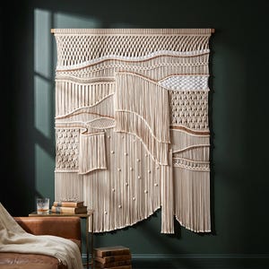 May include: A large, cream-colored macrame wall hanging with a variety of woven textures and patterns. The wall art is mounted on a wooden dowel and hangs against a dark green wall. The piece features fringe and knotted details.