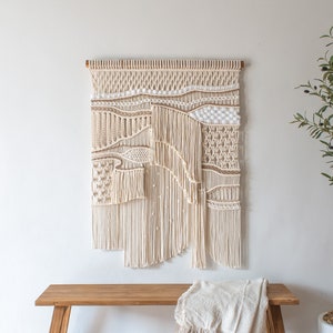 May include: A large macrame wall hanging with a natural beige color. The wall hanging is made of different textures and patterns, creating a unique and modern design. It is hanging on a white wall above a wooden bench with a white throw blanket.