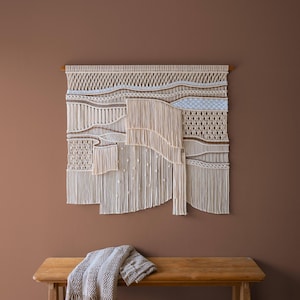 May include: A large, beige macrame wall hanging with a wooden dowel. The textile art piece features various woven patterns and textures, including fringe and knotted details. A wooden bench with a gray blanket with fringe is below the wall hanging.