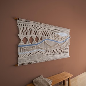 May include: A large macrame wall hanging with a cream-coloured design and a blue accent. The rectangular piece features various woven patterns and is mounted on a wooden dowel. A wooden bench and a blanket are below the wall hanging.