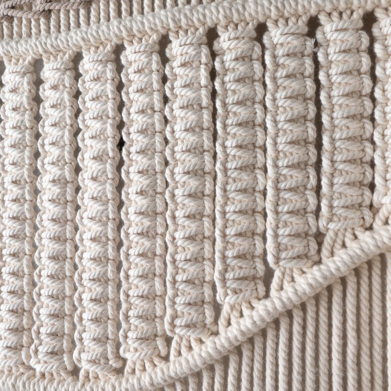 Detailed macro shot of a boho macrame tapestry highlighting the intricate knotting technique. The natural off-white cotton cord is tightly woven into structured, repeating vertical rows, showing expert handmade craftsmanship.