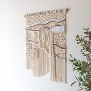 May include: A large, beige macrame wall hanging with a woven pattern and fringe. The hanging is made with natural fibers and has a bohemian style.