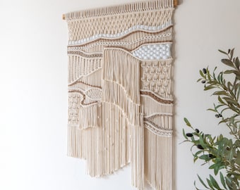 May include: A large, beige macrame wall hanging with a woven pattern and fringe. The hanging is made with natural fibers and has a bohemian style.