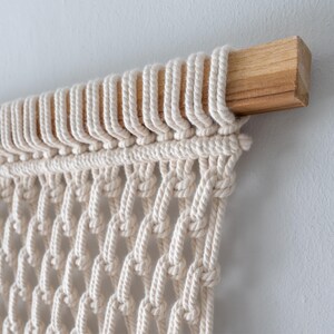 Top detail of a macrame wall art piece showing natural off-white cotton cords cleanly and securely attached to a sturdy, square wooden dowel using uniform lark's head knots.