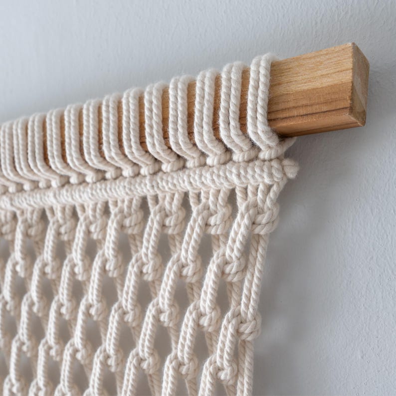Top detail of a macrame wall art piece showing natural off-white cotton cords cleanly and securely attached to a sturdy, square wooden dowel using uniform lark's head knots.
