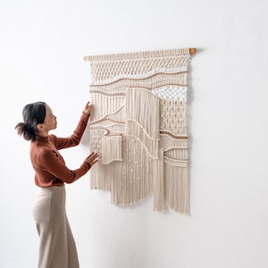 Lifestyle shot of a woman in a rust-colored top adjusting a beautifully textured, handmade boho macrame wall hanging. The image highlights the 3D details, layered fringe, and craftsmanship of the fiber art.