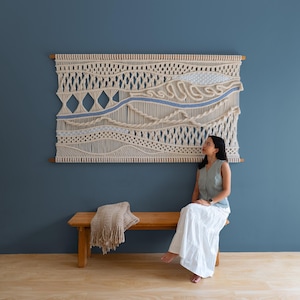May include: A large macrame wall hanging with a natural, cream-colored design and a blue accent. The artwork is mounted on a wooden frame and hangs on a dark blue wall. A woman sits on a wooden bench below the artwork.