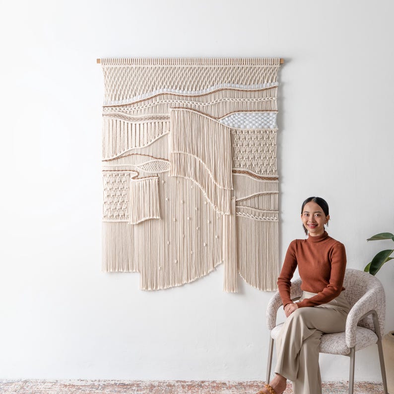A smiling woman sits in a textured modern accent chair next to a large, intricately detailed macrame wall hanging. The minimalist boho living room setting showcases how the fiber art warms up a clean white wall.