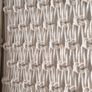 Close-up view of handmade macrame wall decor featuring a classic alternating square knot pattern. The natural cotton cords create an elegant, open netting design within the fiber art piece.