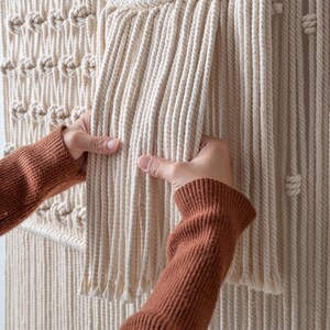 Close-up of a person's hands gently touching the thick, cascading vertical fringe of a handmade macrame wall hanging. The image highlights the high quality, soft texture, and density of the natural cotton cord.