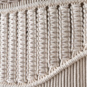 Detailed macro shot of a boho macrame tapestry highlighting the intricate knotting technique. The natural off-white cotton cord is tightly woven into structured, repeating vertical rows, showing expert handmade craftsmanship.