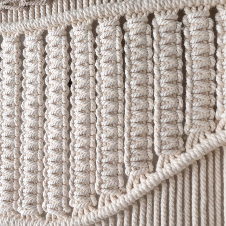 Detailed macro shot of a boho macrame tapestry highlighting the intricate knotting technique. The natural off-white cotton cord is tightly woven into structured, repeating vertical rows, showing expert handmade craftsmanship.