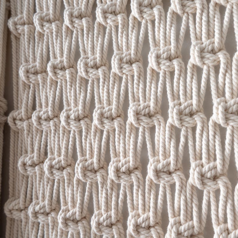 Close-up view of handmade macrame wall decor featuring a classic alternating square knot pattern. The natural cotton cords create an elegant, open netting design within the fiber art piece.