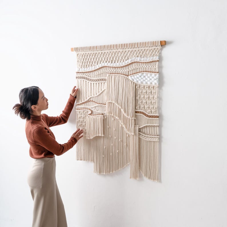 Lifestyle shot of a woman in a rust-colored top adjusting a beautifully textured, handmade boho macrame wall hanging. The image highlights the 3D details, layered fringe, and craftsmanship of the fiber art.