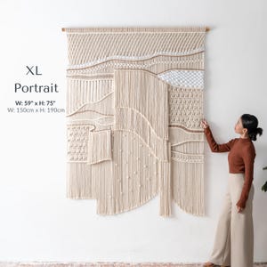 Extra large, tall portrait macrame wall hanging measuring 59 by 75 inches. A woman is standing beside the oversized boho statement piece, highlighting its massive scale, intricate knotting techniques, and floor-length fringe.