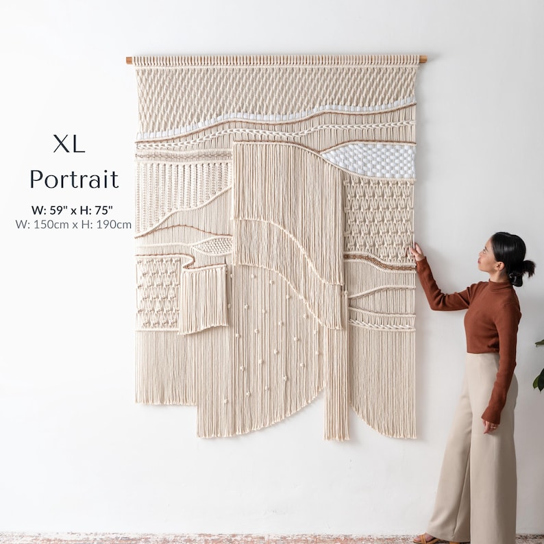 Extra large, tall portrait macrame wall hanging measuring 59 by 75 inches. A woman is standing beside the oversized boho statement piece, highlighting its massive scale, intricate knotting techniques, and floor-length fringe.