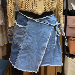 May include: A tan, open-weave cardigan with three large buttons is paired with a denim wrap skirt. The skirt has a frayed hem and a tie closure. The outfit is displayed on a mannequin, with clothing visible in the background.