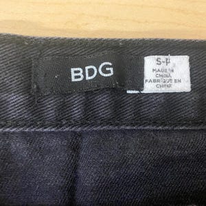 Puede incluir: Primer plano de tejido vaquero negro con costura. Una etiqueta negra dice "BDG" y una etiqueta blanca dice "MADE IN CHINA FABRIQUE EN CHINE".