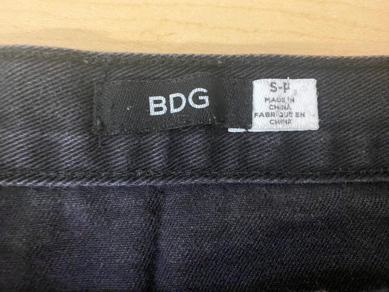 Puede incluir: Primer plano de tejido vaquero negro con costura. Una etiqueta negra dice "BDG" y una etiqueta blanca dice "MADE IN CHINA FABRIQUE EN CHINE".