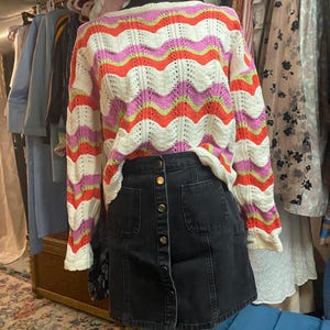 Puede incluir: Un su&eacute;ter colorido con estampado y una falda vaquera negra. El su&eacute;ter presenta un dise&ntilde;o ondulado en blanco, naranja, rosa y verde. La falda tiene una botonadura frontal y dos bolsillos delanteros. El conjunto se exhibe en un maniqu&iacute;.