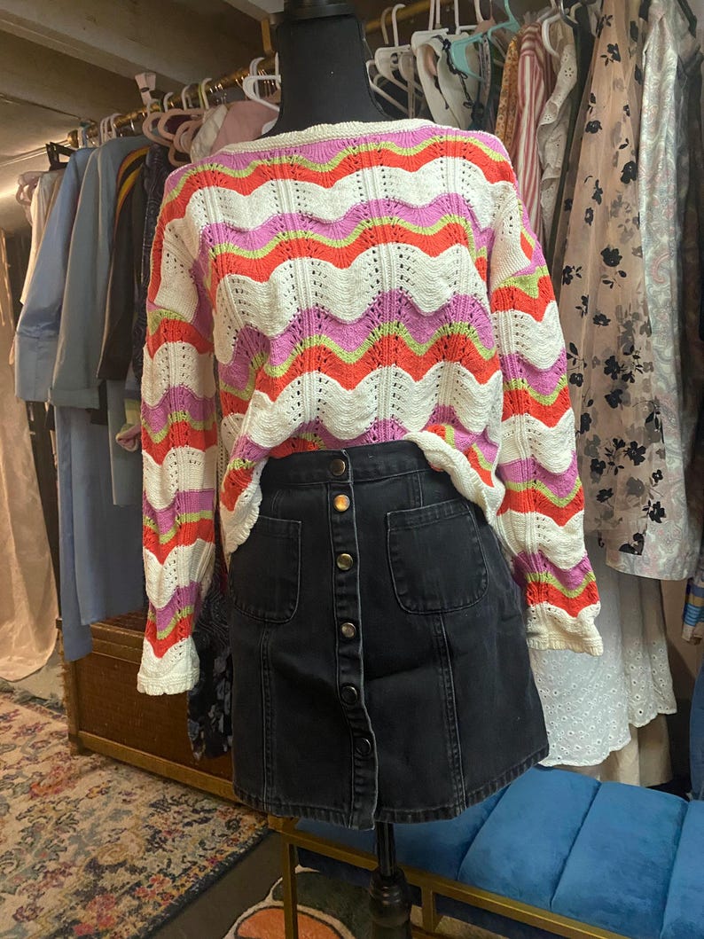 Puede incluir: Un su&eacute;ter colorido con estampado y una falda vaquera negra. El su&eacute;ter presenta un dise&ntilde;o ondulado en blanco, naranja, rosa y verde. La falda tiene una botonadura frontal y dos bolsillos delanteros. El conjunto se exhibe en un maniqu&iacute;.