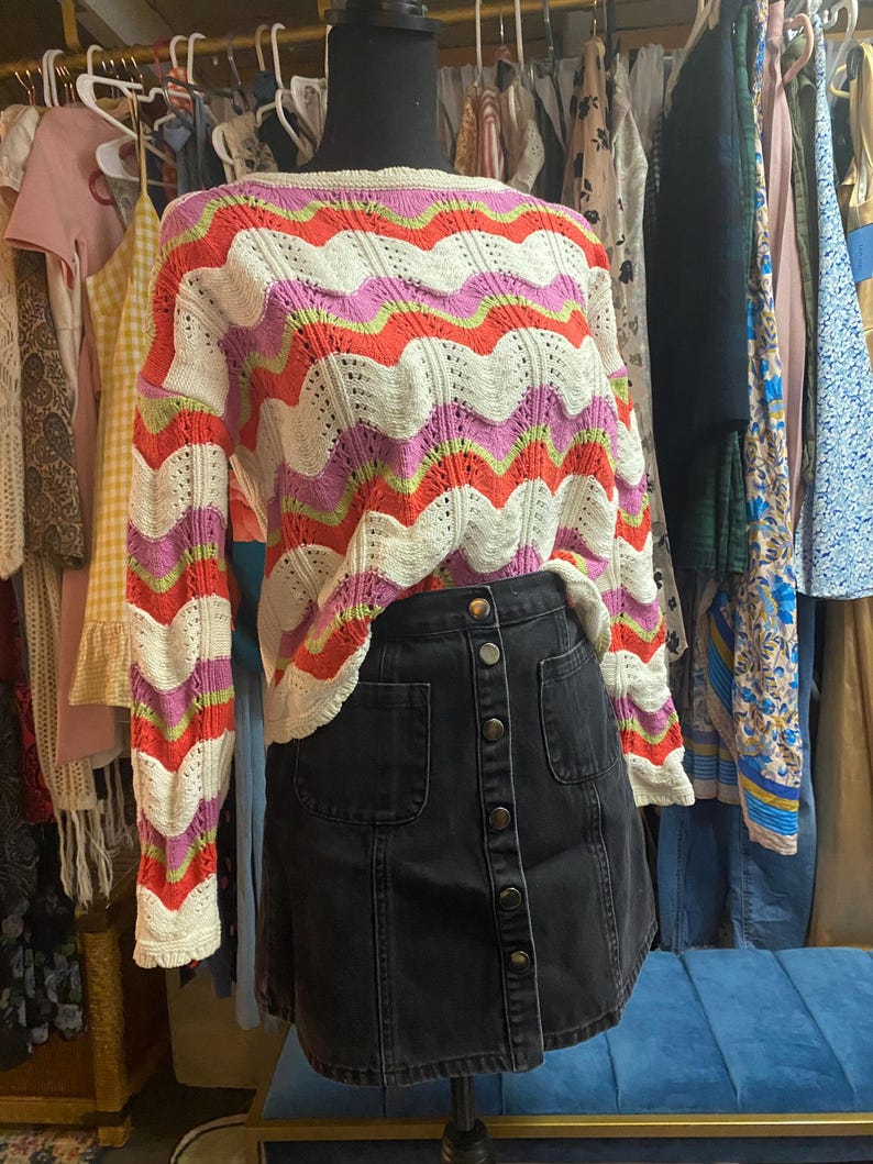 Puede incluir: Un maniqu&iacute; exhibe un su&eacute;ter colorido y una falda de mezclilla. El su&eacute;ter presenta un patr&oacute;n de ondas en blanco, rosa, naranja y verde. La falda es de mezclilla de lavado oscuro con botones frontales y dos bolsillos delanteros. Ropa.