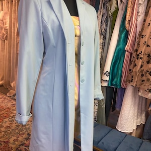 May include: A light blue blazer-style dress with a three-button closure and long sleeves. The dress is displayed on a mannequin, showcasing its tailored fit. A variety of other clothing items hang in the background.