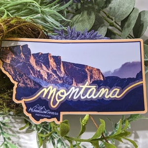 Pode incluir: Um autocolante em forma de Montana, com uma paisagem montanhosa com a palavra "montana" em escrita cursiva. O autocolante também inclui o texto "Alpenglow Studio Photography". O autocolante é rodeado por vegetação.