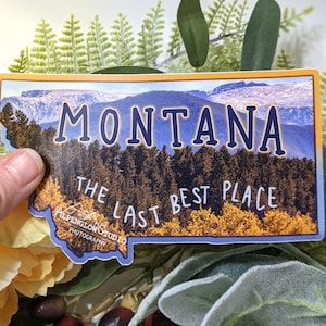 Pode incluir: Um adesivo azul e amarelo com o contorno do estado de Montana. O adesivo apresenta uma imagem cênica de montanhas e árvores com o texto "MONTANA THE LAST BEST PLACE" e o logotipo "ALPENGLOW STUDIO PHOTOGRAPHY".