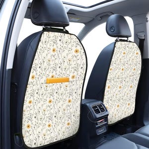 May include: Car seat covers featuring a floral design. The covers are cream-coloured with a repeating pattern of yellow flowers and grey stems. They have a black border and a pocket on the back.