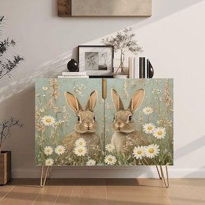 May include: A cabinet with a light blue background featuring two rabbits surrounded by daisies and wildflowers. The cabinet has gold hairpin legs and a wooden top. The artwork has a vintage, spring-like aesthetic.