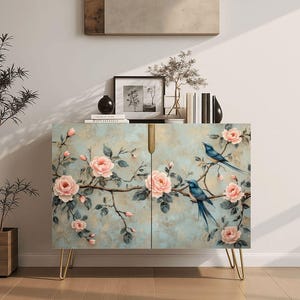 May include: A two-door cabinet with a floral and bird design. The cabinet features a light blue background with pink roses and blue birds. It has gold-colored hairpin legs and a gold-colored handle.