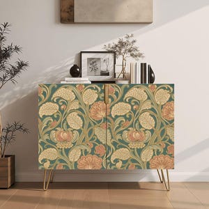 May include: A rectangular cabinet featuring two doors with a repeating floral pattern in shades of muted green, cream, and soft peach. The cabinet is supported by four thin, gold-toned metal hairpin legs and includes a vertical gold-toned handle.