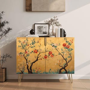 May include: A rectangular cabinet with a gold-toned finish featuring a painted design of dark tree branches with red and pink flowers and green leaves. The cabinet has two doors with a vertical handle and is supported by four thin gold metal legs.
