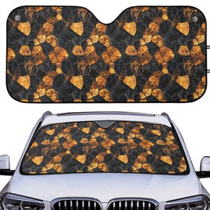 May include: A car windshield sunshade with a black and gold stone pattern. The sunshade is rectangular with cutouts for the rearview mirror and is shown installed in a white car. The pattern is repeated on the sunshade.
