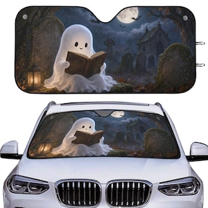 May include: A rectangular car windshield sun shade featuring a graphic illustration of a white ghost sitting in a graveyard at night, reading an open book under a full moon. The scene includes tombstones, a stone mausoleum, and a glowing lantern.