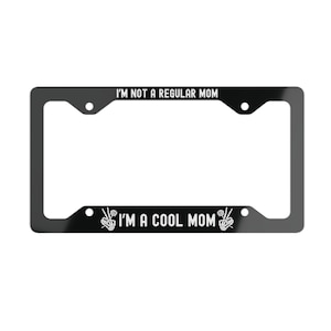 May include: Black license plate frame with the text "I'M NOT A REGULAR MOM" at the top and "I'M A COOL MOM" at the bottom. The phrase is in white, with peace sign graphics.