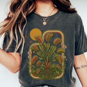 May include: A charcoal gray short-sleeve t-shirt featuring a graphic print of a Venus flytrap plant with green leaves and red-rimmed traps, surrounded by small white flowers and a yellow sun, framed by a rounded rectangular border with orange lines.