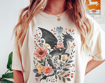 May include: A cream-colored t-shirt with a black dragon surrounded by colorful flowers. The dragon has blue wings and a pink flower on its head. The text "ZEN & SHAPE" is printed on the bottom right corner of the shirt.