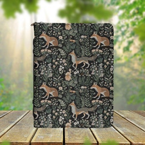 Könnte beinhalten: Ein schwarzer Notizbuchumschlag mit einem sich wiederholenden Muster aus Füchsen und floralen Designs. Die Füchse sind orange und weiß, und die Blumen sind in Schattierungen von Rosa, Lila und Grün.