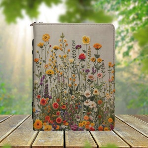 Könnte beinhalten: Ein beigefarbener Leinen-Notizbuchumschlag mit einem gestickten Blumenmuster. Die Blumen sind in den Farben Gelb, Orange, Rot und Lila gehalten. Der Notizbuchumschlag ist perfekt, um Ihrem Alltag einen Hauch von Natur zu verleihen.