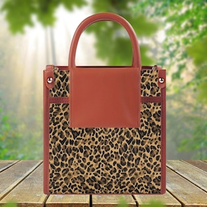 May include: A structured handbag featuring a leopard print pattern on the main body and a solid terracotta-colored top handle and front flap. The bag has silver-toned hardware accents on the sides and the text ZEN & SHAPE in the bottom corner.