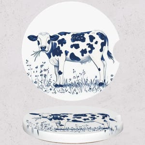May include: White car coasters with a blue and white illustration of a cow grazing in a field. The cow has black spots and is eating grass. The coasters are round with a small indentation for easy removal.