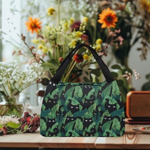 May include: A black lunch bag with a green tropical leaf and black cat print. The bag has a black handle and a zipper closure.